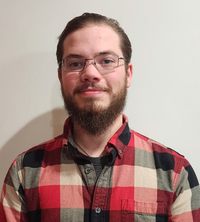 Portrait photo of white male-presenting individual with facial hair. They are wearing glasses and a red flannel shirt.