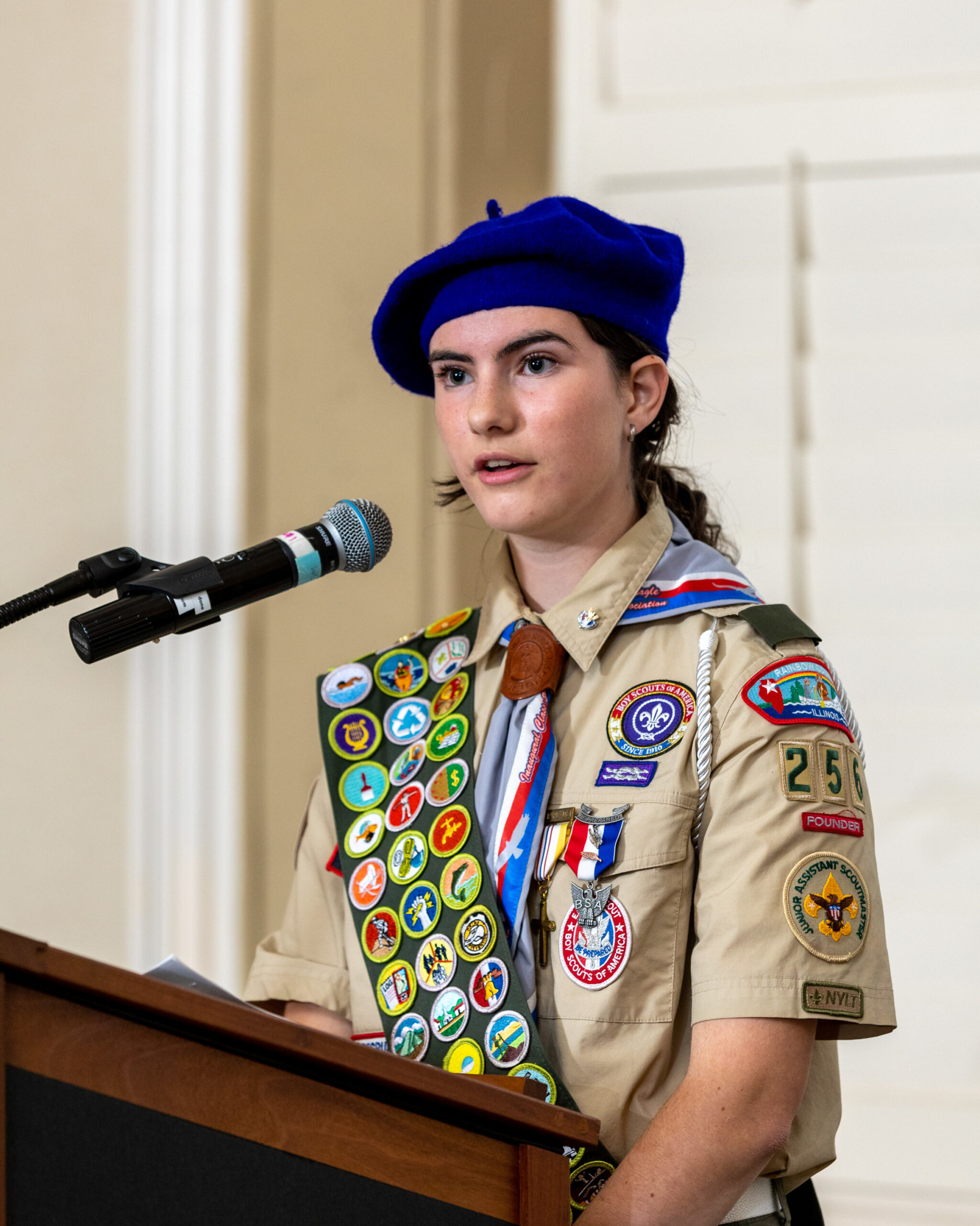 Portrait photo of female-presenting individual with long dark hair. They are wearing a tan scouting uniform with several badges and a hat. They are standing and speaking at a podium.