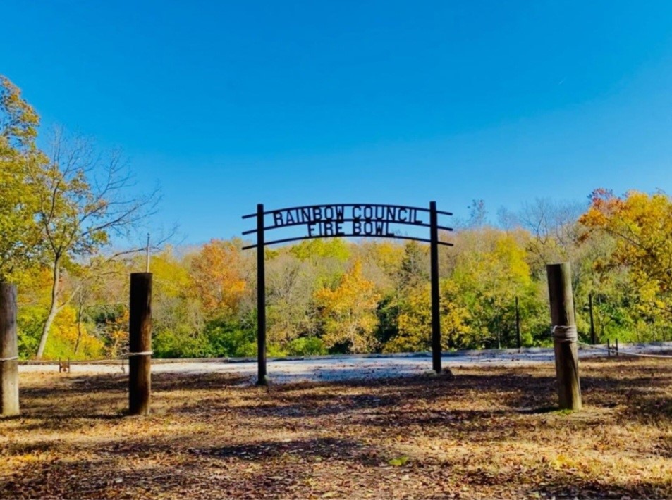 Landscape photo of the woods at Rainbow Scout Reservation. There are two posts with rope, and a large sigh in the back reading "Rainbow Council Fire Bowl"