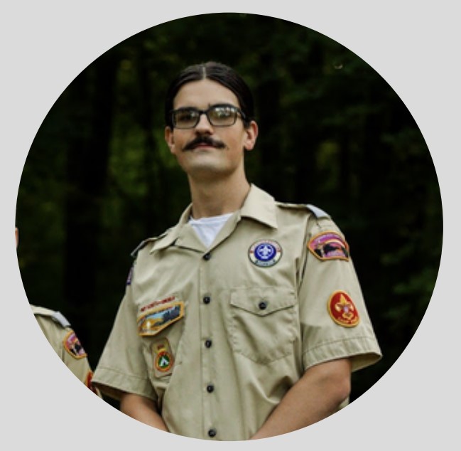 Portrait photo of male-presenting individual with a very pronounced moustache and dark hair. They are wearing a tan scouting uniform with several badges and embroidery reading "Boy Scouts of America"