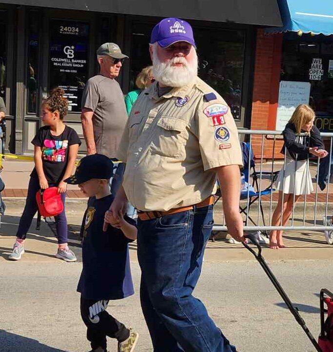 Photo of a male-presenting scout youth and male-presenting scout adult holding hands and pulling a cart, walking on a main street. Youth is wearing a lions cub tee and adult is wearing a scouting uniform shirt with several badges and a RSR baseball cap. There are bystanders in the background watching the procession.
