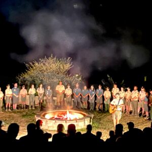 Photo of a group of scouts, at night, standing around a fire bowl. There are both male and female-presenting young adults. There is a male-presenting individual holding a guitar, walking around the fire bowl. There is a cloud of smoke coming from the fire bowl, billowing up, into the night.