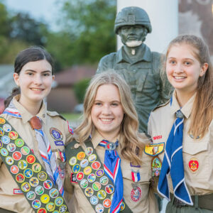 A group photo of female presenting individuals, one with brunette hair, two with blonde hair, of various heights, standing in front of a memorial statue. All three are wearing tan scout uniforms, with several badges and kerchiefs.