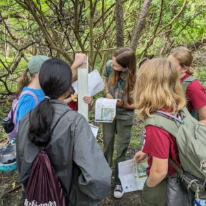 Group of female-presenting youths standing around a trail marker in the middle of the woods. They are wearing casual hiking clothes and backpacks. They are holding maps and comparing information.