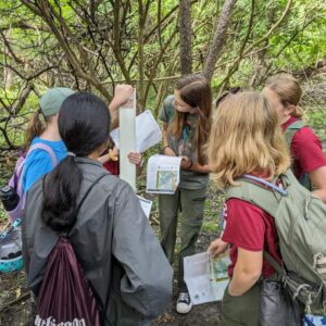 Group of female-presenting youths standing around a trail marker in the middle of the woods. They are wearing casual hiking clothes and backpacks. They are holding maps and comparing information.