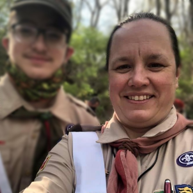 Portrait of two people, one heavily blurred, the other a female-presenting adult standing in a forest. They are both wearing scouting uniforms and smiling for a selfie.