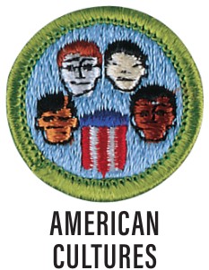 Image of the American Cultures merit badge. The badge is blue with a green border with the faces of diverse youth around a flag.
