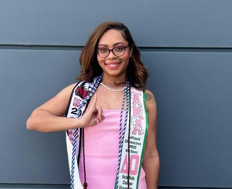 A black, female presenting person posting for a photo while wearing graduation regalia. She is wearing a pink dress, white strappy sandals, glasses and doing a sorority hand signal.