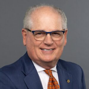 Portrait of male presenting person in a blue business suit. They are wearing glasses and an orange tie.