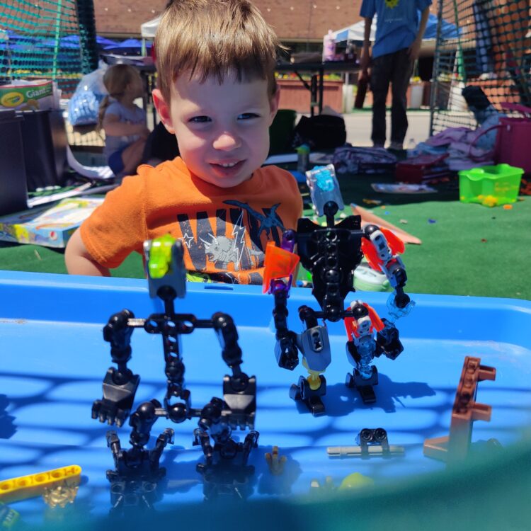 Photo of a youth standing in front of an activity table. There are robots in front of the youth and the youth is smiling.