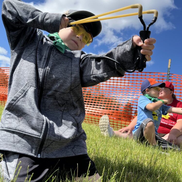 Photo of a child in a field, wearing safety goggles,, holding a slingshot.